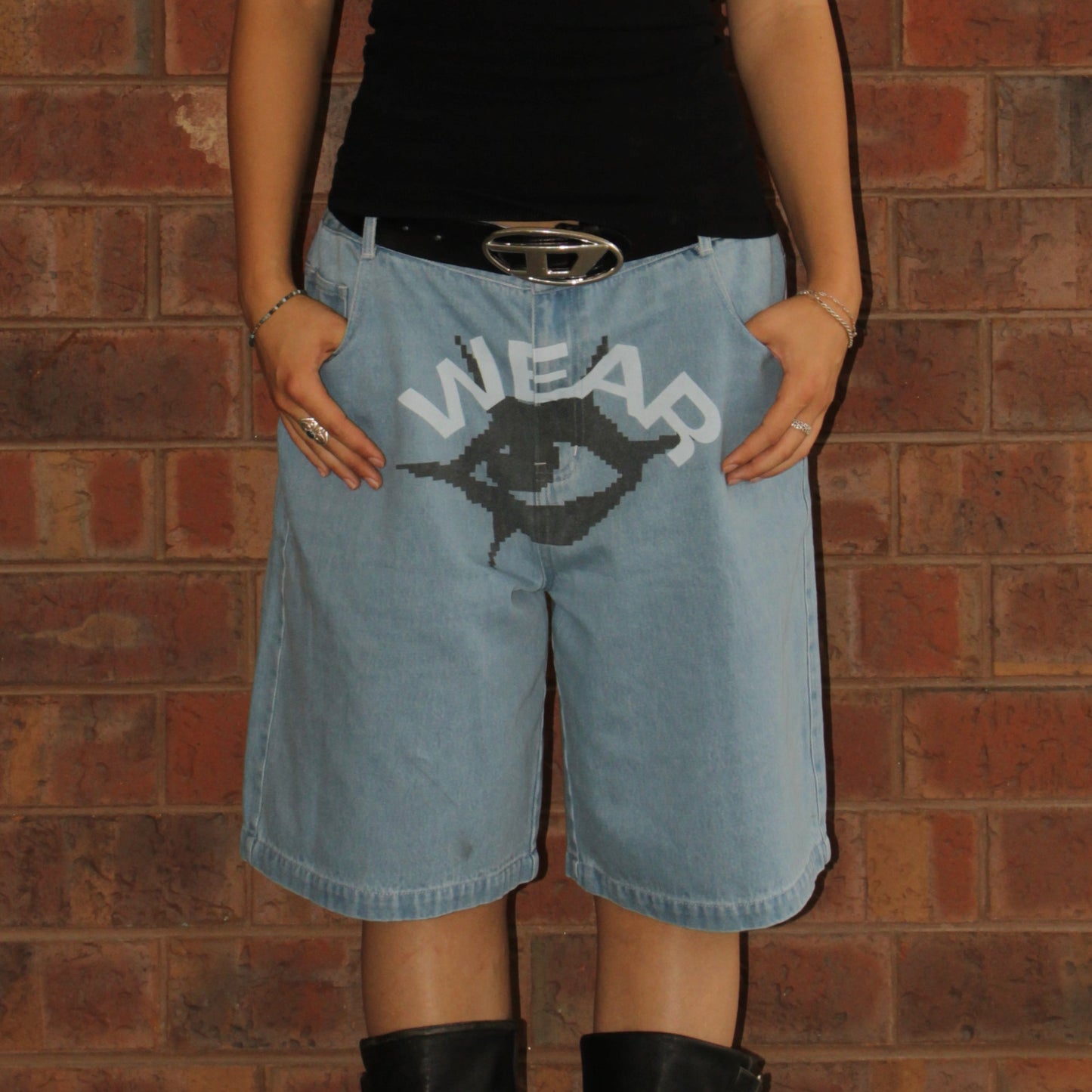 Person wearing light blue denim shorts with a graphic design and text, standing against a brick wall.