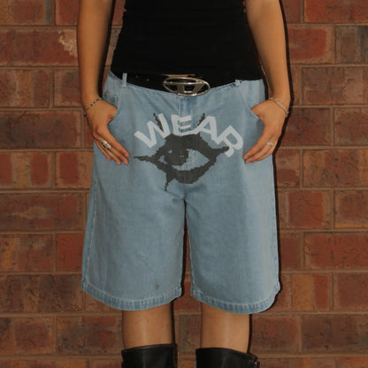 Person wearing light blue denim shorts with a graphic design and text, standing against a brick wall.